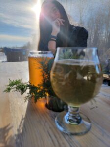 a glass of cider and a glass of beer rest on a table. the sun is shining in the background and a woman is smiling slightly.