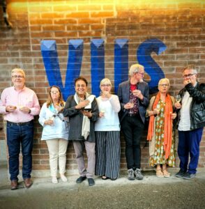 six guests stand outside vij's restaurant in Vancouver. Celebrity chef Vikram Vij is in the middle of them. They are all holding a glass of champagne and tasting some street food.