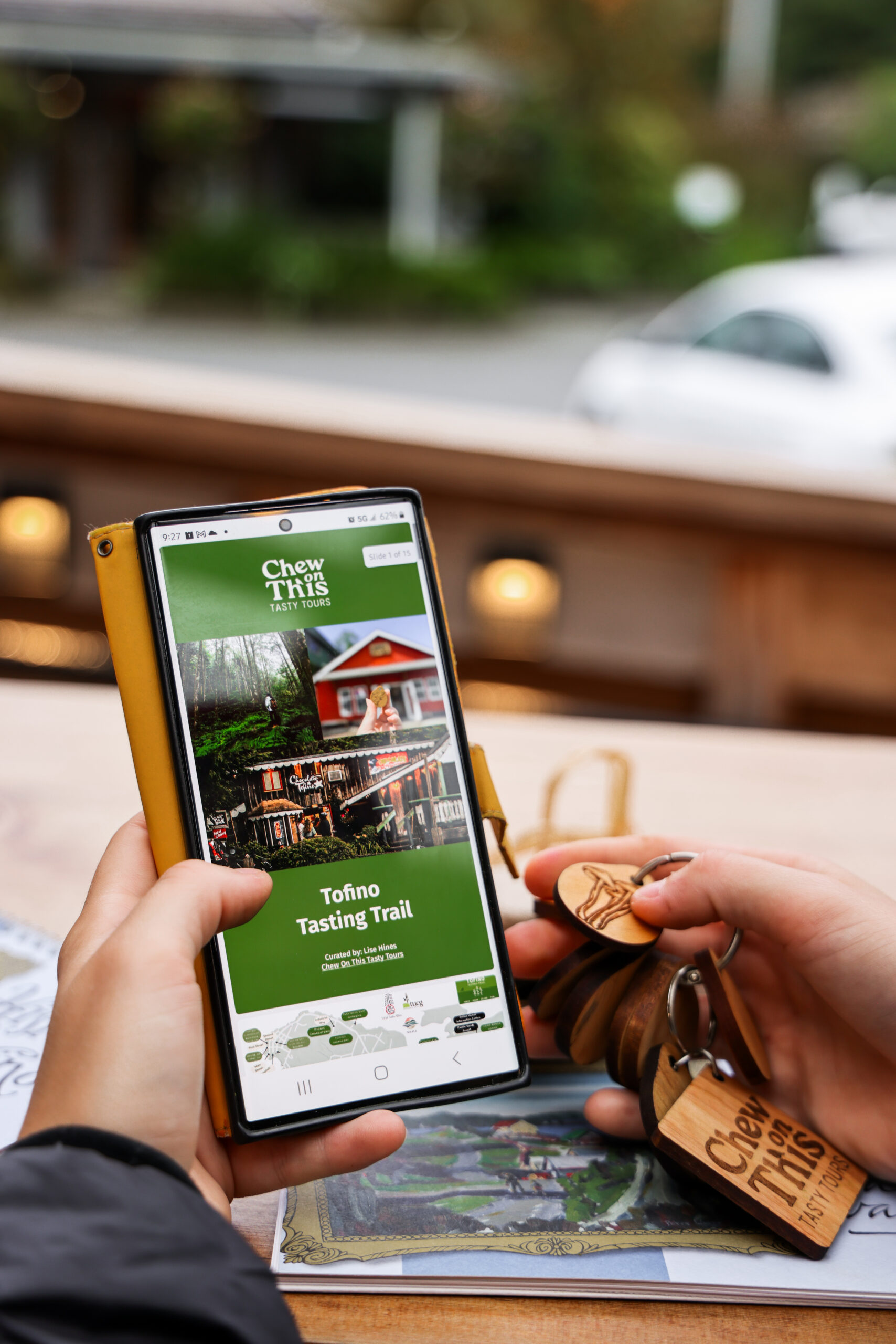 a person holding a cell phone looking at an itinerary for the Tofino Tasting Trail Encounters tour. In the other hand there are wooden tokens with engravings on them