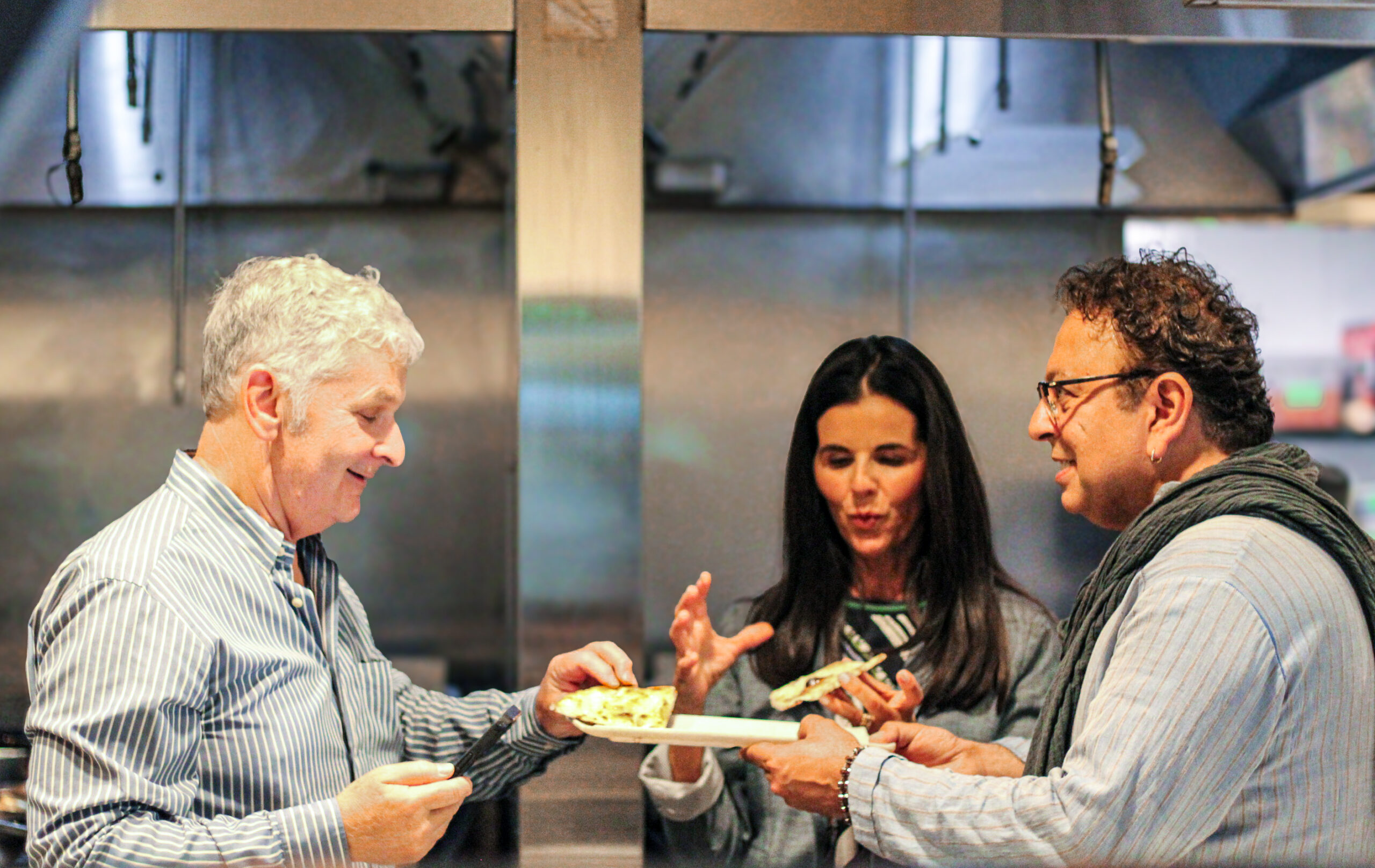 two people standing in a restaurant kitchen interacting with chef vikram vij who is offering them a taste of food.