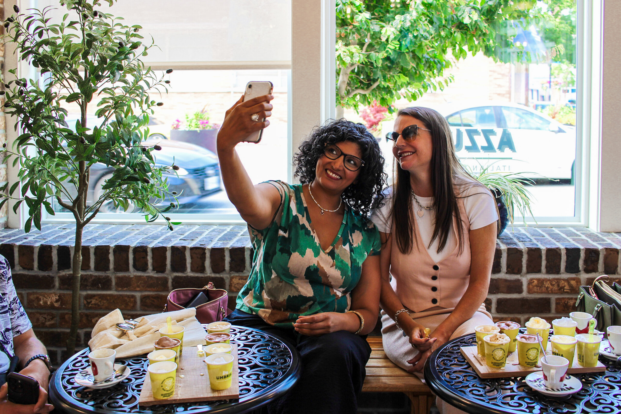 two women with food on the table in front of them take a selfie. both are smiling. one has a mobile phone raised to capture a photo of herself.