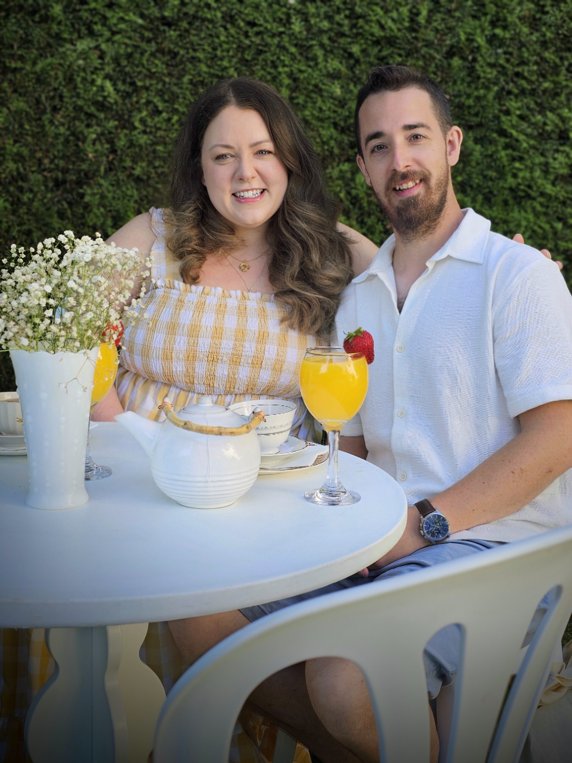 a couple is sitting at a white table in a garden. they have drinks in front of them and they are smiling into the camera.